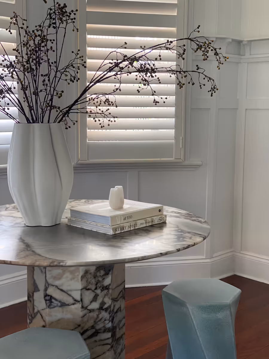 Round marble table holding a sculptural vase and stacked books, beside plantation shutters