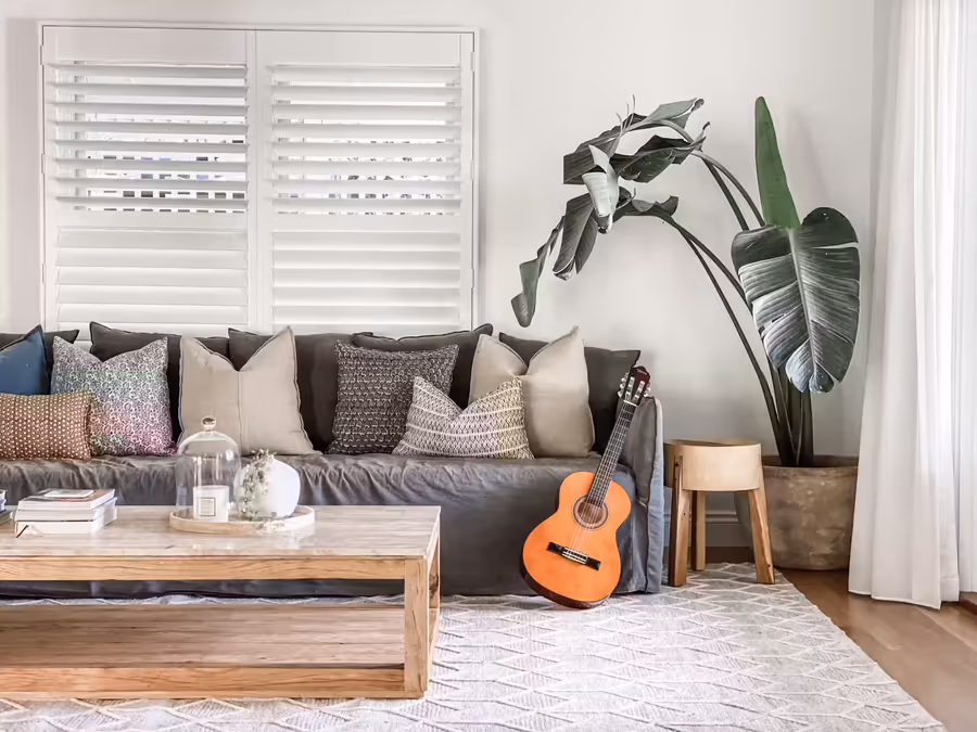 Slouchy grey sofa below plantation shutters, with a guitar and potted greenery alongside