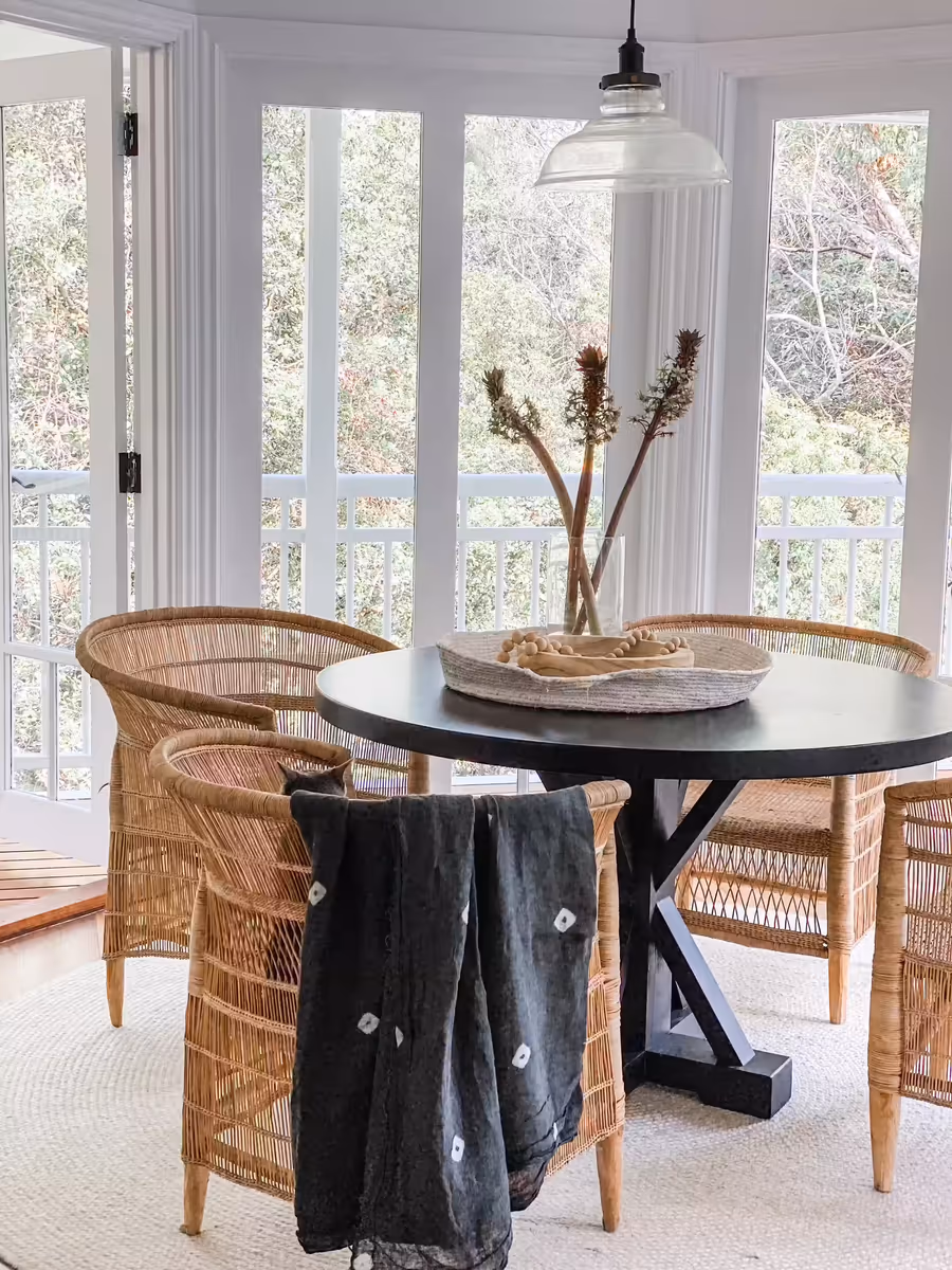 Round dining table and woven rattan chairs in the window bay overlooking the bush