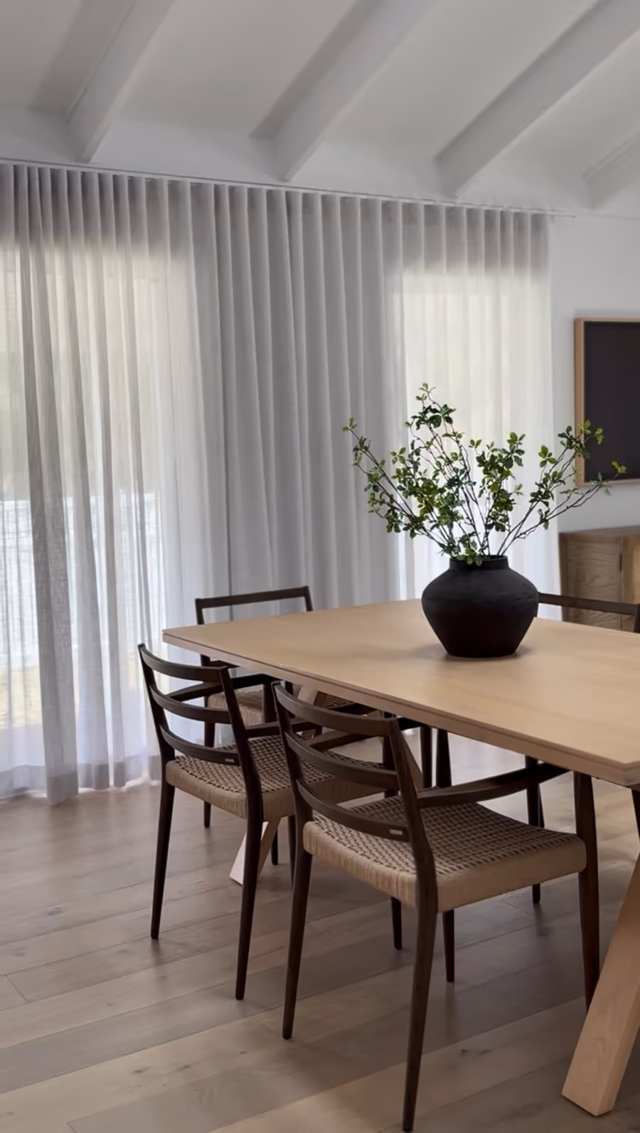 Oak dining table and woven-seat chairs against sheer curtains under a raked ceiling