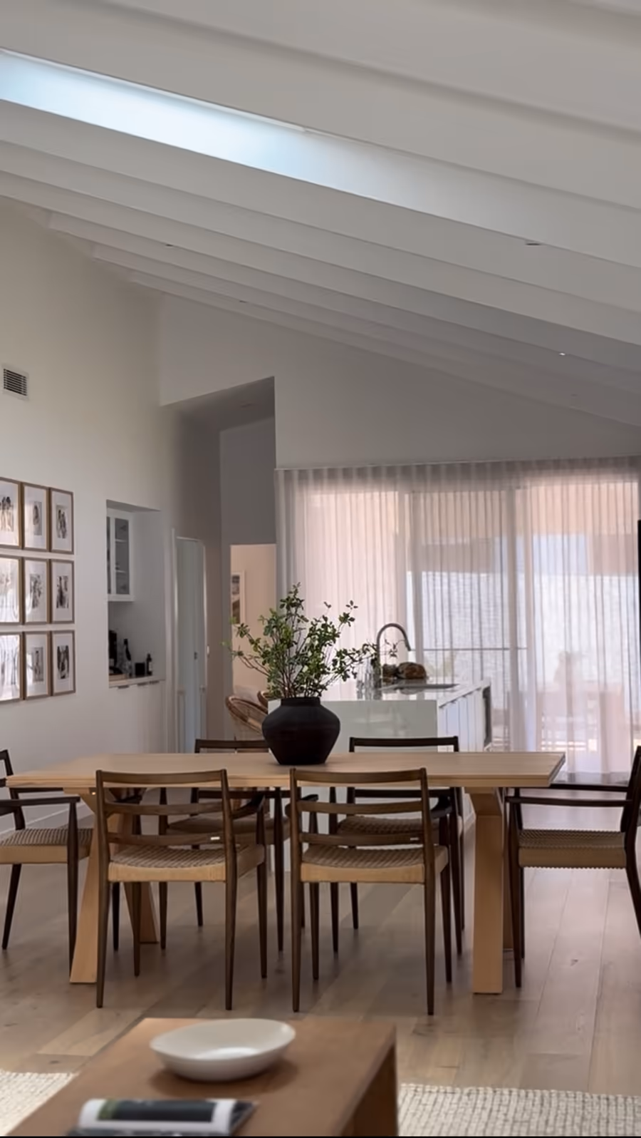 Raked ceiling over the dining table, with the kitchen and sheer-curtained wall beyond