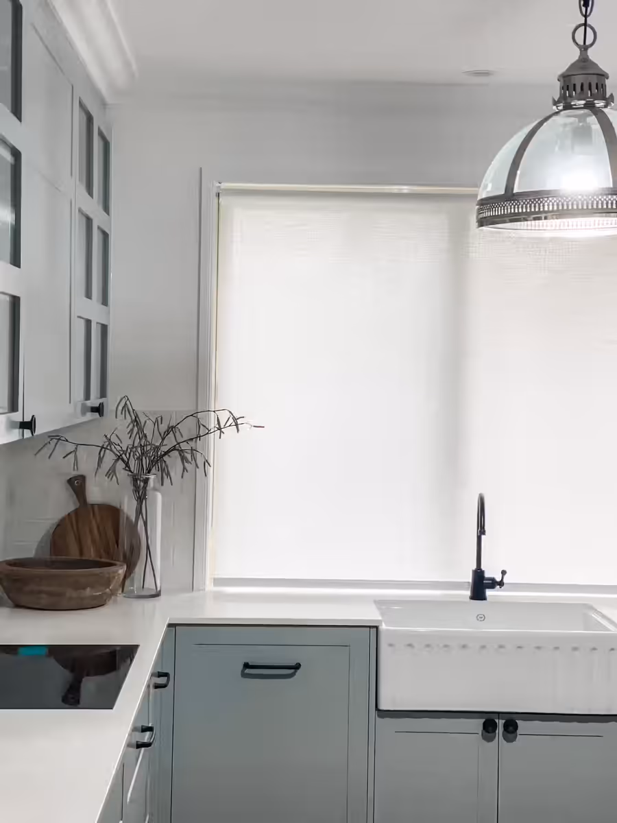 Farmhouse sink and stone benchtop below a roller blind, with a black tapware