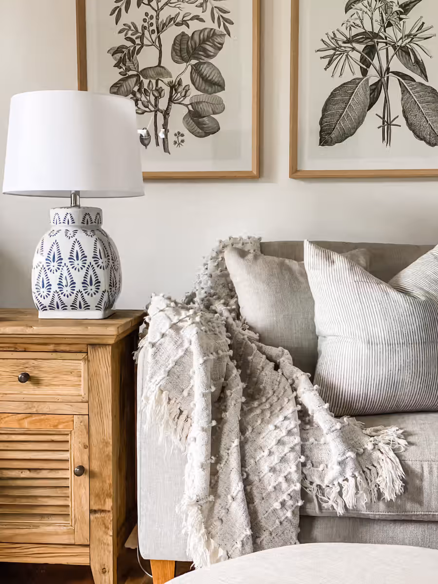 Botanical prints above a grey sofa, with a patterned lamp on a timber side cabinet