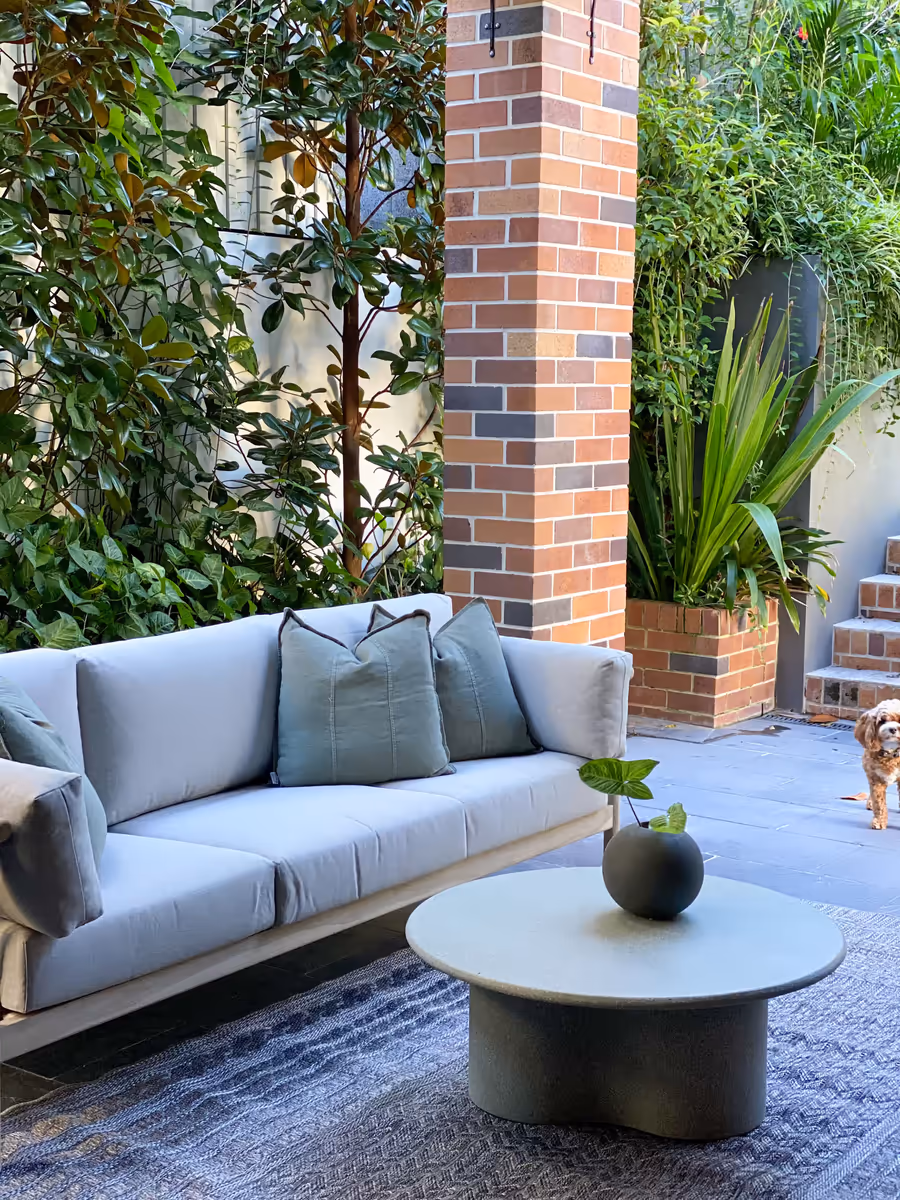 Pale outdoor sofa and a dark round table on the courtyard, backed by a green wall