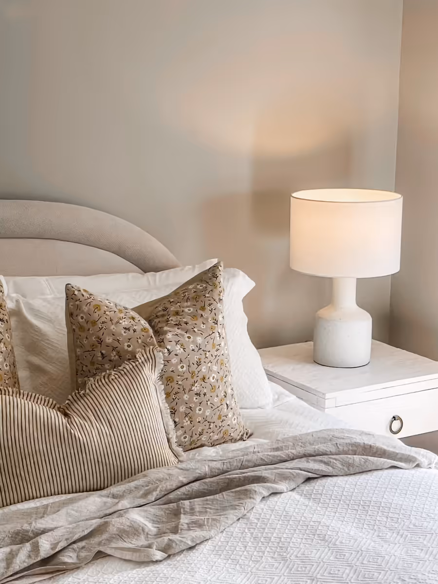 Curved bedhead with floral and striped cushions, beside a white ceramic table lamp