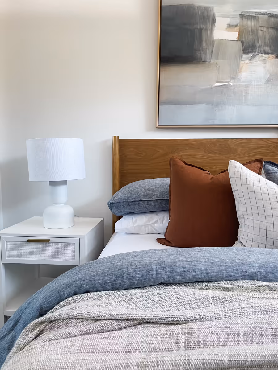 Timber bedhead with rust and denim bedding below a grey abstract canvas