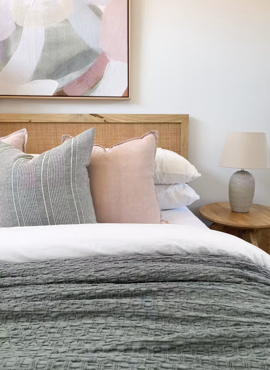 Rattan bedhead with blush and striped cushions, a ceramic lamp alongside