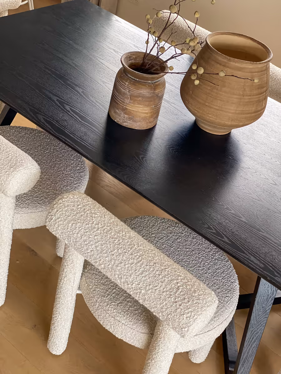 Boucle dining chairs tucked under a black timber table holding two turned vessels