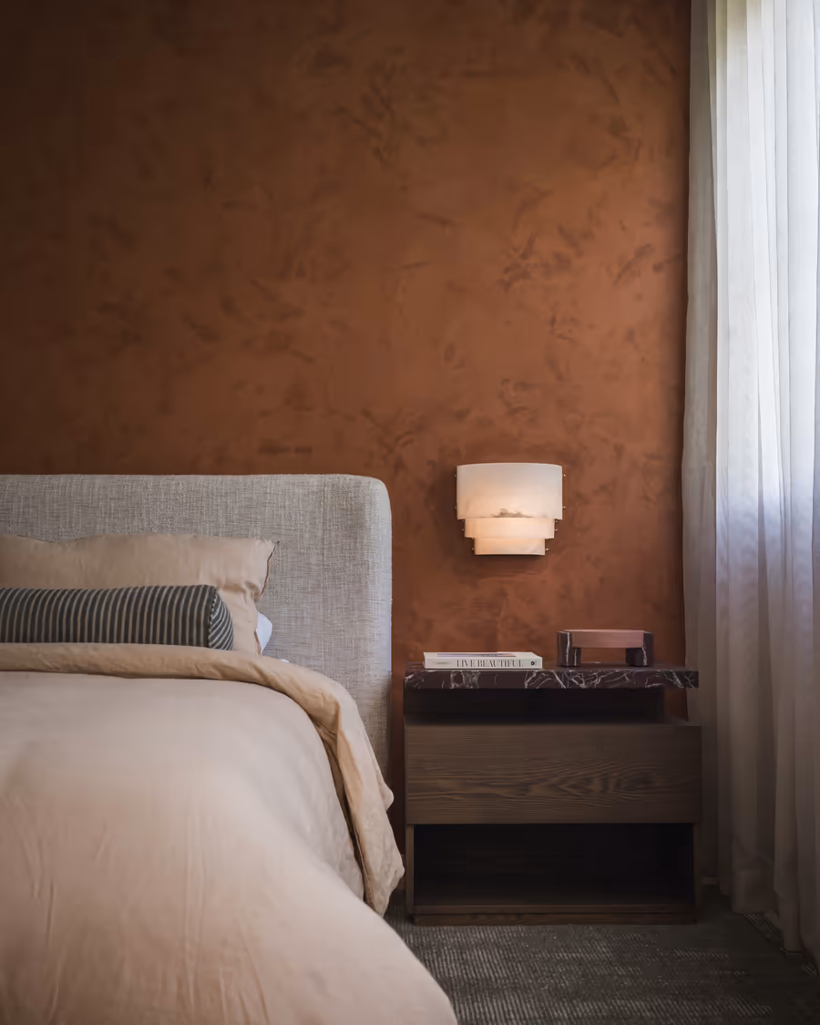 Upholstered bedhead against a limewashed rust wall, lit by an alabaster wall light