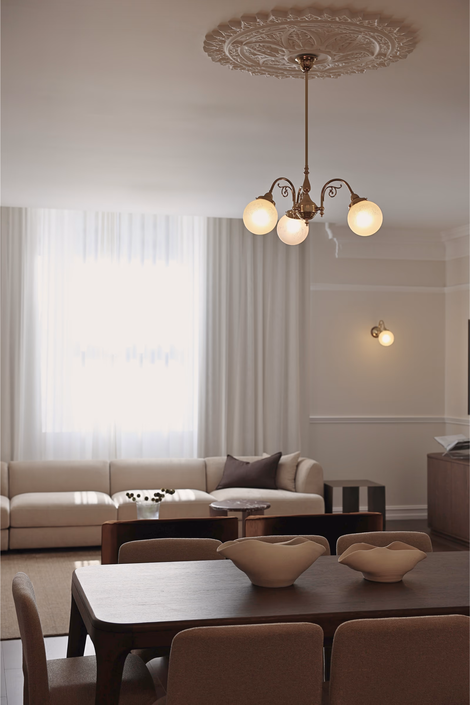 Living and dining under a heritage ceiling rose and brass chandelier, sheers at the window