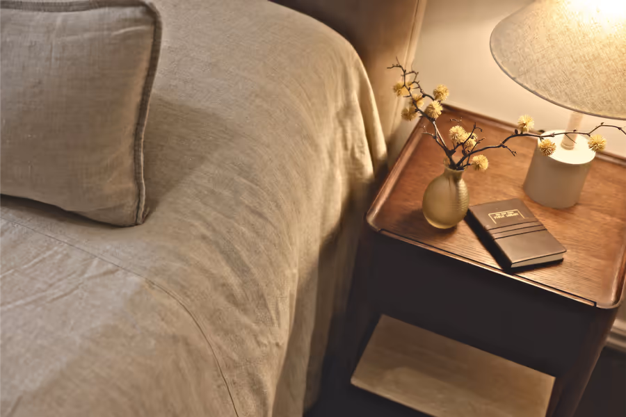 Linen bed with a lamp and dried stems on the dark timber bedside table