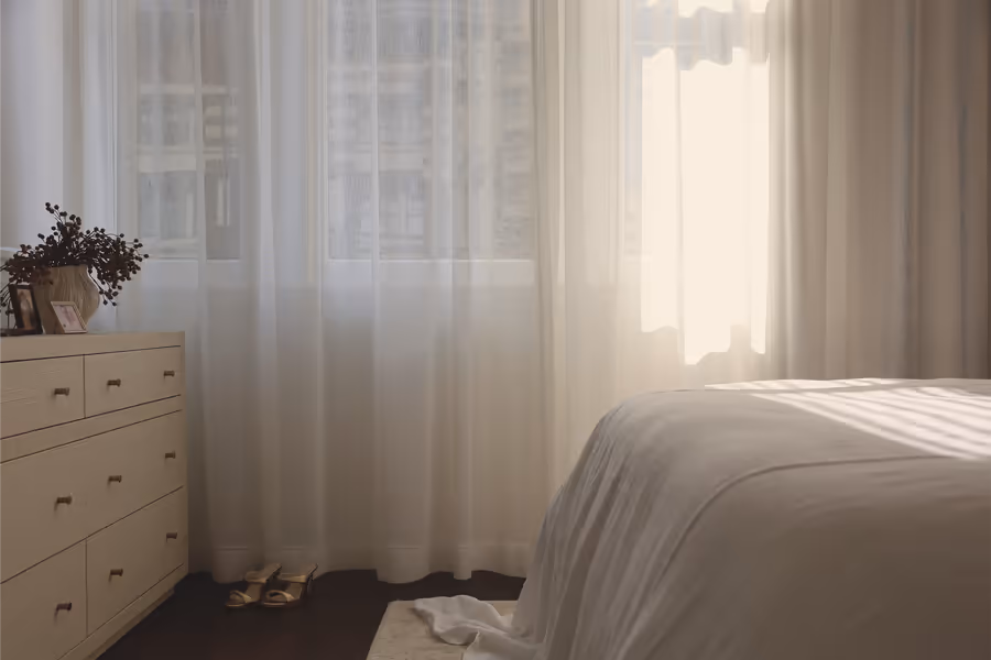 Dressing chest beside full-length sheers, with the bed catching the afternoon light