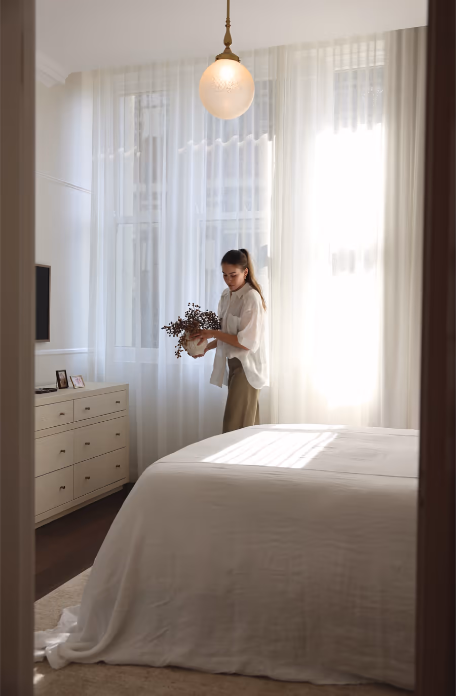 Carrying dried stems past the bed, beneath a frosted glass pendant