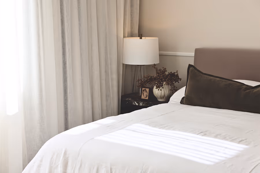 White linen bedding and a brown bolster, with a lamp on the dark bedside table